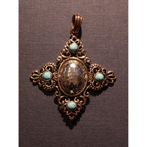 Pode incluir: Pingente dourado com um design de cruz ornamentado. O pingente apresenta um centro oval prateado martelado e quatro pedras de cor turquesa. Os braços da cruz têm volutas decorativas. O pingente tem uma argola para uma corrente.