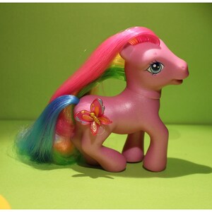 May include: A pink toy pony with a rainbow mane and tail. A butterfly-shaped accessory is attached to its side. The pony has blue eyes and is standing on a green surface.