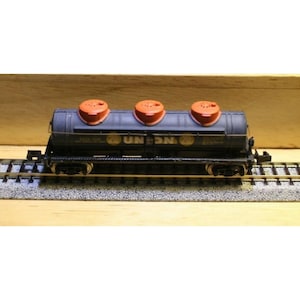 May include: A model train tank car in dark gray with three orange circular hatches on top. The side of the cylindrical tank features the text UNION in yellow lettering, flanked by two circular logos. The car is mounted on black wheels.