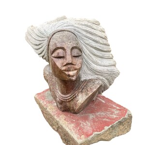 Puede incluir: Una escultura de piedra de un busto de mujer con los ojos cerrados y cabello suelto. La escultura es una mezcla de tonos marrones y grises, descansando sobre una base rectangular roja y gris. La obra de arte tiene una expresión serena y contemplativa.
