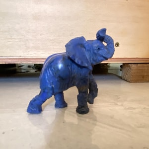 May include: A blue elephant figurine with its trunk raised. The elephant has large ears and is walking forward. The figurine is made of a solid material and is sitting on a reflective surface.