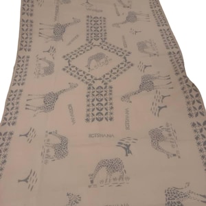 May include: A beige textile with a repeating pattern of giraffes and trees in grey. The word "Botswana" is printed in grey. The design includes geometric shapes and borders, creating a safari theme. The textile is likely cotton or linen.