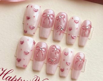 Puede incluir: Un conjunto de uñas postizas rosas claras decoradas con corazones y lazos, perfectas para el Día de San Valentín. Las uñas se muestran en una tarjeta con el mensaje "Happy Valentine's Day, My Love". Cintas rosas y rojas rodean las uñas.