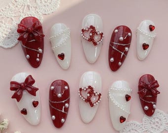 Puede incluir: Un conjunto de diez uñas artificiales con varios diseños. Algunas uñas son blancas con perlas y detalles de corazón, mientras que otras son rojas con lazos, perlas y detalles de corazón. Las uñas están dispuestas sobre una superficie rosa con encaje.