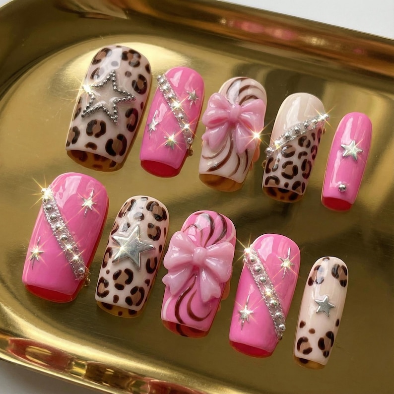 K&ouml;nnte beinhalten: Eine Sammlung von zehn k&uuml;nstlichen Fingern&auml;geln in verschiedenen Designs. Die N&auml;gel zeigen rosa, Leopardenmuster und Schleifenmotive mit silbernen Sternen und Strasssteinen. Die N&auml;gel werden auf einer goldfarbenen Oberfl&auml;che pr&auml;sentiert.