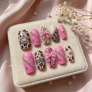K&ouml;nnte beinhalten: Ein Set von zehn k&uuml;nstlichen N&auml;geln in verschiedenen Designs. Die N&auml;gel weisen rosa, Leopardenmuster und cremefarbene Farben auf. Einige N&auml;gel haben silberne Sterne und Strasssteine, w&auml;hrend andere Schleifendetails aufweisen. Die N&auml;gel werden auf einer wei&szlig;en Samtoberfl&auml;che pr&auml;sentiert.