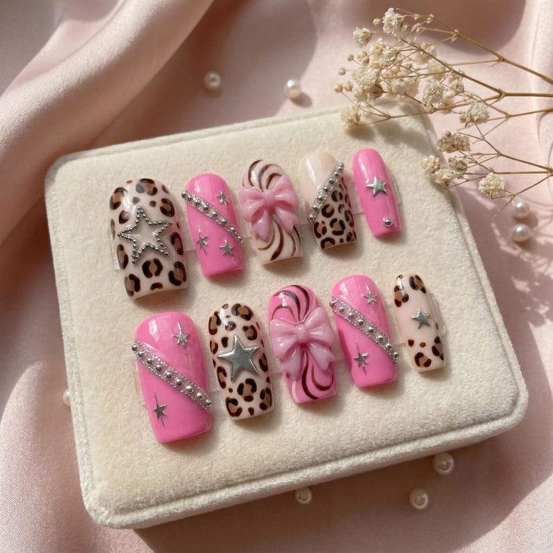 K&ouml;nnte beinhalten: Ein Set von zehn k&uuml;nstlichen N&auml;geln in verschiedenen Designs. Die N&auml;gel weisen rosa, Leopardenmuster und cremefarbene Farben auf. Einige N&auml;gel haben silberne Sterne und Strasssteine, w&auml;hrend andere Schleifendetails aufweisen. Die N&auml;gel werden auf einer wei&szlig;en Samtoberfl&auml;che pr&auml;sentiert.