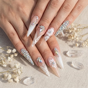 May include: Long, pointed acrylic nails with various designs. Some nails are adorned with sparkling rhinestones, others with delicate white floral accents. Several nails feature intricate white lace patterns, creating a glamorous and elegant look.