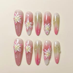 May include: A set of ten artificial nails with a gradient design, transitioning from pink to white and yellow. Each nail features a 3D flower with white petals and yellow centers, and some have small, clear droplet accents.