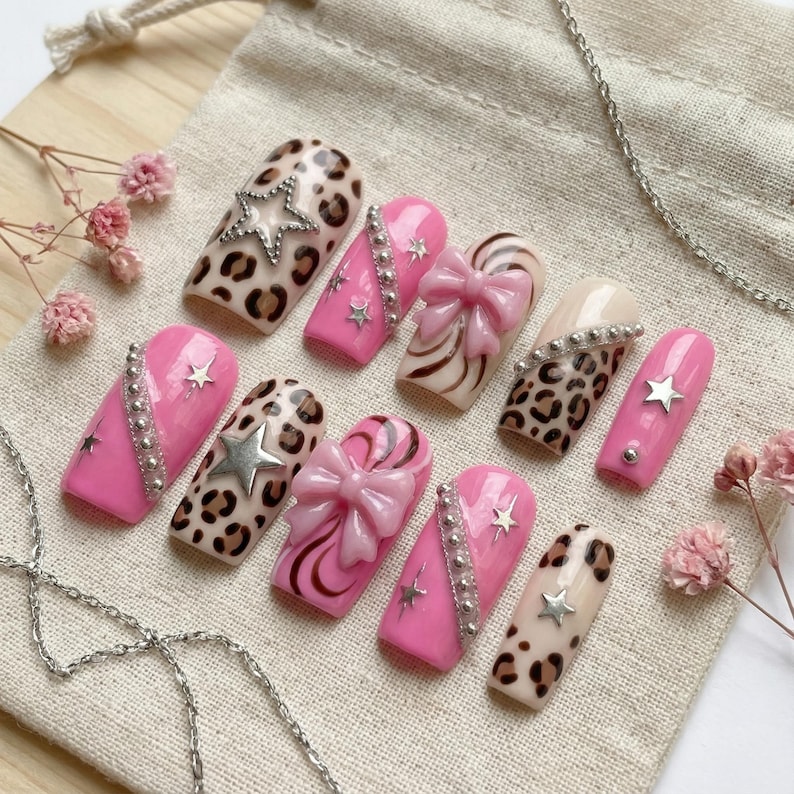 K&ouml;nnte beinhalten: Eine Sammlung k&uuml;nstlicher Fingern&auml;gel mit verschiedenen Designs. Einige N&auml;gel haben eine rosa Basis mit silbernen Akzenten und Sternverzierungen. Andere zeigen einen Leopardenmusterdruck, w&auml;hrend einige Schleifendetails aufweisen. Die N&auml;gel sind auf einer neutralen Stoffoberfl&auml;che angeordnet.