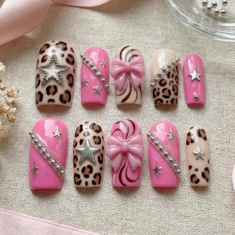 K&ouml;nnte beinhalten: Ein Set aus zehn k&uuml;nstlichen N&auml;geln in verschiedenen Designs. Die N&auml;gel weisen rosa, Leopardenmuster und Schleifendesigns auf. Einige N&auml;gel haben silberne Stern- und Strassakzente. Die N&auml;gel sind auf einer neutralen Stoffoberfl&auml;che angeordnet.