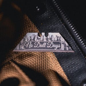 May include: A silver-toned utility knife blade featuring a laser-etched pattern of fish along the top edge and the text "No Dull Days" in a cursive script centered on the metal surface, resting against a textured brown and black fabric background.