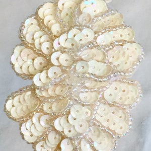 Pode incluir: Sequins brancos iridescentes e contas são costurados juntos em um padrão de flor. Os sequins são dispostos em espiral, criando um efeito tridimensional.