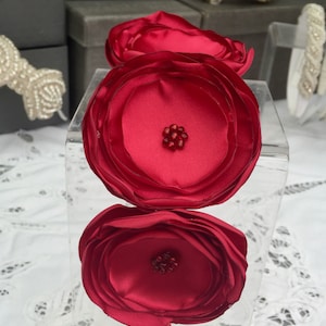 Könnte beinhalten: Ein rotes Satin-Haarband mit drei großen, übereinanderliegenden Stoffblumen. Jede Blume hat einen kleinen roten Strassstein-Cluster in der Mitte.