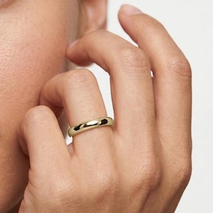 Könnte beinhalten: Ein goldener Bandring mit poliertem Finish. Der Ring hat ein schlichtes, klassisches Design, wahrscheinlich aus Gold. Der Ring befindet sich an einem Finger, was darauf hindeutet, dass er getragen wird. Die glatte Oberfläche des Rings reflektiert das Licht und hebt seine Form hervor.
