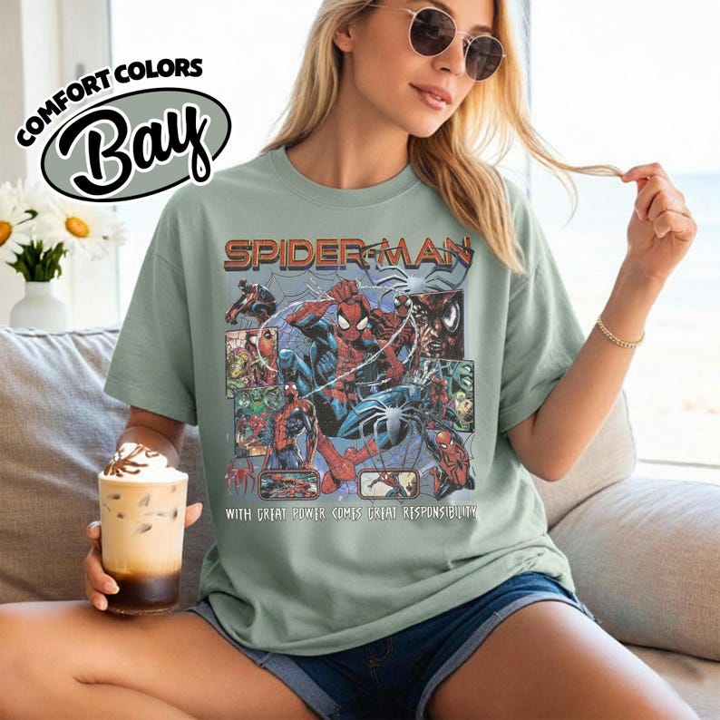 K&ouml;nnte beinhalten: Ein salbeigr&uuml;nes T-Shirt mit einem Spider-Man-Motiv. Das Design zeigt Comic-Panels mit Spider-Man in Action. Der Text "SPIDER-MAN" befindet sich &uuml;ber der Grafik, und "WITH GREAT POWER COMES GREAT RESPONSIBILITY" darunter.