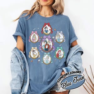 May include: Blue jean colored t-shirt featuring oval portraits of animated princesses. Each portrait is framed with a decorative bow. The shirt has the words "COMFORT COLORS" and "Blue Jean" printed on the bottom right.