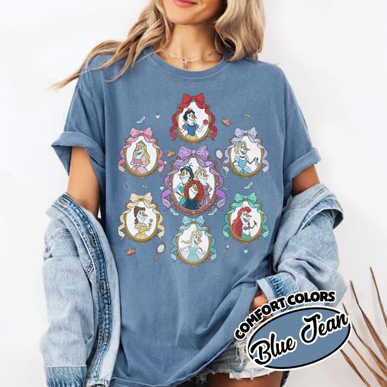 May include: Blue jean colored t-shirt featuring oval portraits of animated princesses. Each portrait is framed with a decorative bow. The shirt has the words "COMFORT COLORS" and "Blue Jean" printed on the bottom right.