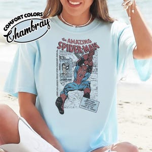 K&ouml;nnte beinhalten: Hellblaues T-Shirt mit einer Comic-Grafik von Spider-Man. Das Design enth&auml;lt den Text "the AMAZING SPIDER-MAN" und "COMFORT COLORS Chambray". Die Grafik zeigt Spider-Man in Aktion, zusammen mit Comic-Panels und Text.