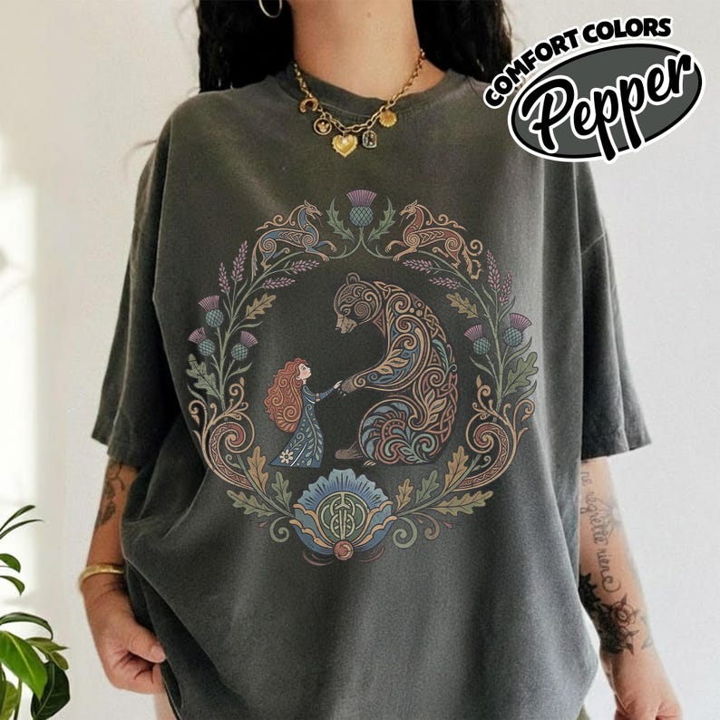 May include: Dark grey t-shirt with a circular graphic featuring a bear and a person holding hands, surrounded by floral and animal designs. The text "COMFORT COLORS Pepper" is printed on the upper right.