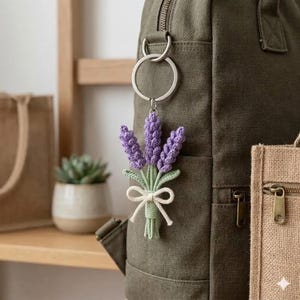 May include: A crocheted lavender bouquet keychain with purple flowers, green leaves, and a white bow. The keychain is attached to a silver ring and hangs from a green backpack. A small succulent in a white pot is visible in the background.