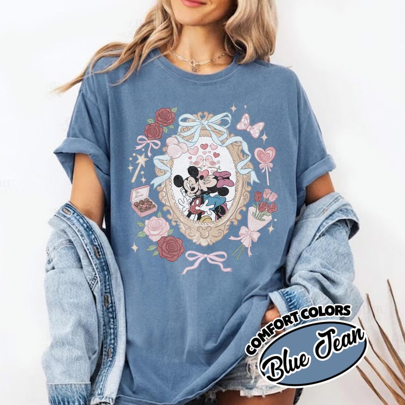 Puede incluir: Una camiseta de color azul jean con un gr&aacute;fico de Mickey y Minnie Mouse dentro de un marco ovalado ornamentado. El dise&ntilde;o incluye rosas rosas y rojas, un lazo, una caja de bombones y piruletas en forma de coraz&oacute;n. El texto COMFORT COLORS BLUE JEAN es visible.