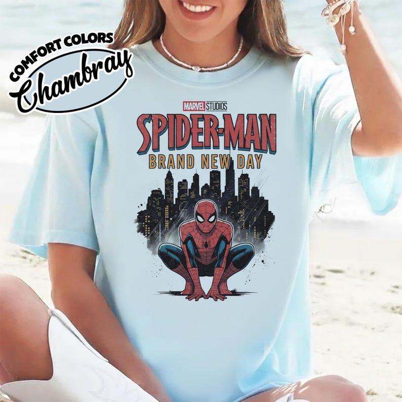 Puede incluir: Una camiseta azul claro con un gr&aacute;fico de Spider-Man agachado frente a un horizonte urbano negro. El texto dice Marvel Studios Spider-Man Brand New Day. La camiseta tambi&eacute;n incluye un logotipo de Comfort Colors Chambray en la esquina.