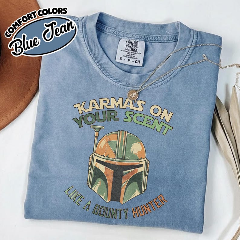 May include: A blue Comfort Colors t-shirt with the text "Karma's on Your Scent Like a Bounty Hunter" and a graphic of a helmet. The shirt has a crew neck and short sleeves. A silver necklace is draped across the shirt.