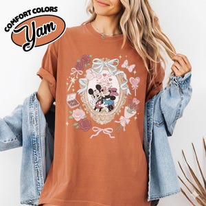 Puede incluir: Una camiseta de manga corta color &oacute;xido con un gr&aacute;fico de Mickey y Minnie Mouse dentro de un marco ovalado ornamentado. El dise&ntilde;o incluye cintas azul claro, rosas rosadas, una caja de bombones, un ramo de flores y corazones peque&ntilde;os.