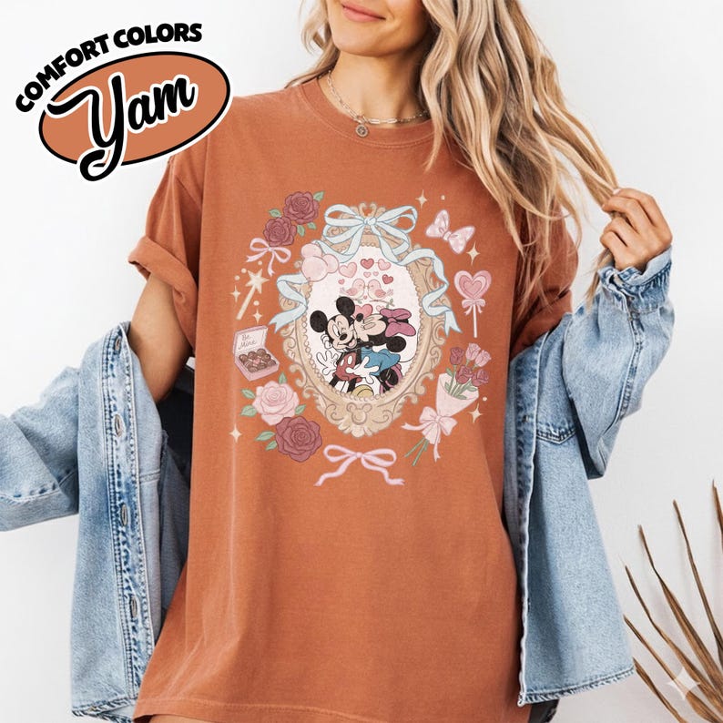 Puede incluir: Una camiseta de manga corta color &oacute;xido con un gr&aacute;fico de Mickey y Minnie Mouse dentro de un marco ovalado ornamentado. El dise&ntilde;o incluye cintas azul claro, rosas rosadas, una caja de bombones, un ramo de flores y corazones peque&ntilde;os.