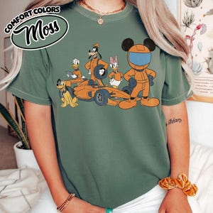 May include: Green t-shirt with a cartoon graphic of Disney characters in racing attire, including Mickey Mouse, Goofy, Donald Duck, and Daisy Duck, with a race car. The text "COMFORT COLORS Moss" is in a green oval on the upper left.