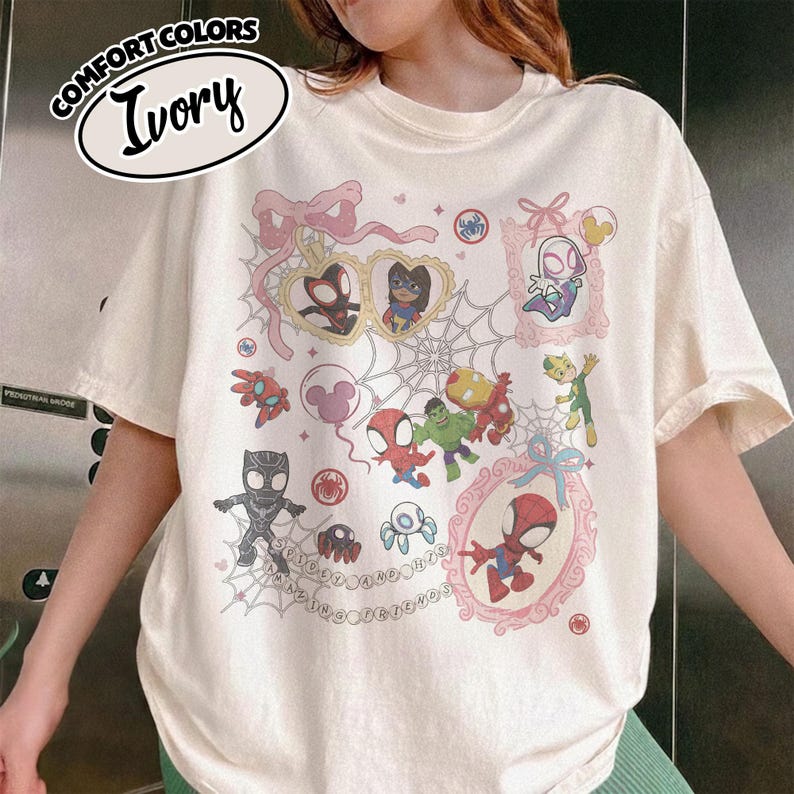 K&ouml;nnte beinhalten: Elfenbeinfarbenes T-Shirt mit einer farbenfrohen Grafik verschiedener Spider-Man-Charaktere und zugeh&ouml;riger Bilder. Das Design umfasst gerahmte Portr&auml;ts, Spinnennetze und die Texte "Comfort Colors" und "Ivory".