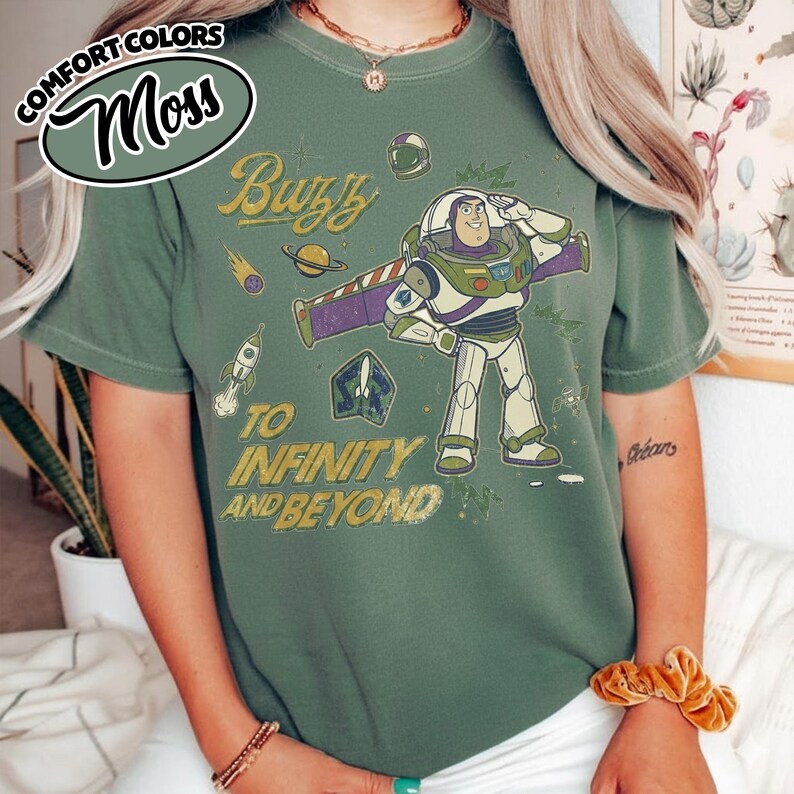 May include: A moss green t-shirt featuring a graphic of Buzz Lightyear saluting, surrounded by space-themed icons including a rocket, planet, and helmet. The text reads Buzz TO INFINITY AND BEYOND in a distressed yellow font across the front.