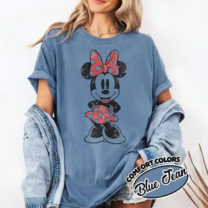 May include: A blue t-shirt with a distressed Minnie Mouse graphic, sporting a red polka dot bow and dress. The sleeves are rolled up. The text "COMFORT COLORS Blue Jean" is in a circular design.