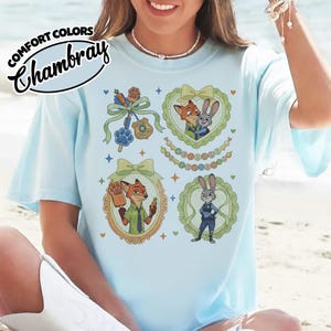 May include: A light blue t-shirt featuring a graphic print of Zootopia characters Nick Wilde and Judy Hopps. The design includes four distinct sections with ornate frames, ribbons, and small stars, alongside the text Comfort Colors Chambray.