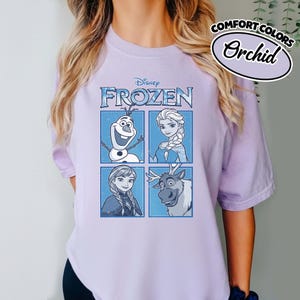 May include: A light purple t-shirt featuring a graphic of characters from Disney's Frozen. The design includes Olaf, Elsa, Anna, and Sven in a four-panel layout. The shirt also has the words "COMFORT COLORS" and "Orchid" printed on it.