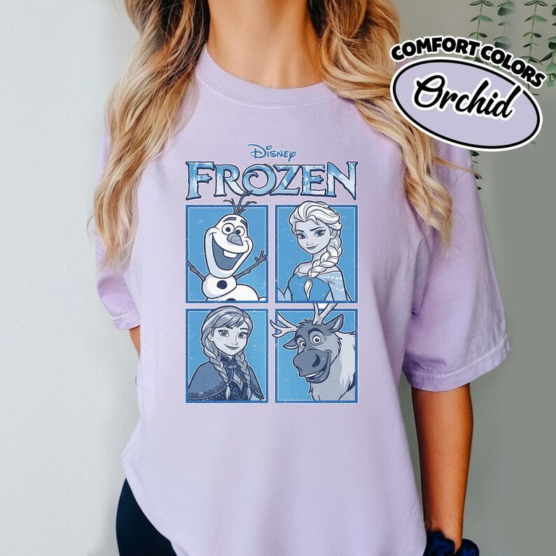 May include: A light purple t-shirt featuring a graphic of characters from Disney's Frozen. The design includes Olaf, Elsa, Anna, and Sven in a four-panel layout. The shirt also has the words "COMFORT COLORS" and "Orchid" printed on it.