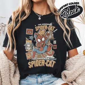 May include: Black t-shirt featuring a cartoon illustration of a cat dressed as Spider-Man, with the text "THE AMEOWZING SPIDER-CAT." The design includes a smaller cat character and various stamp-like graphics. The words "COMFORT COLORS" and "Black" are also visible.
