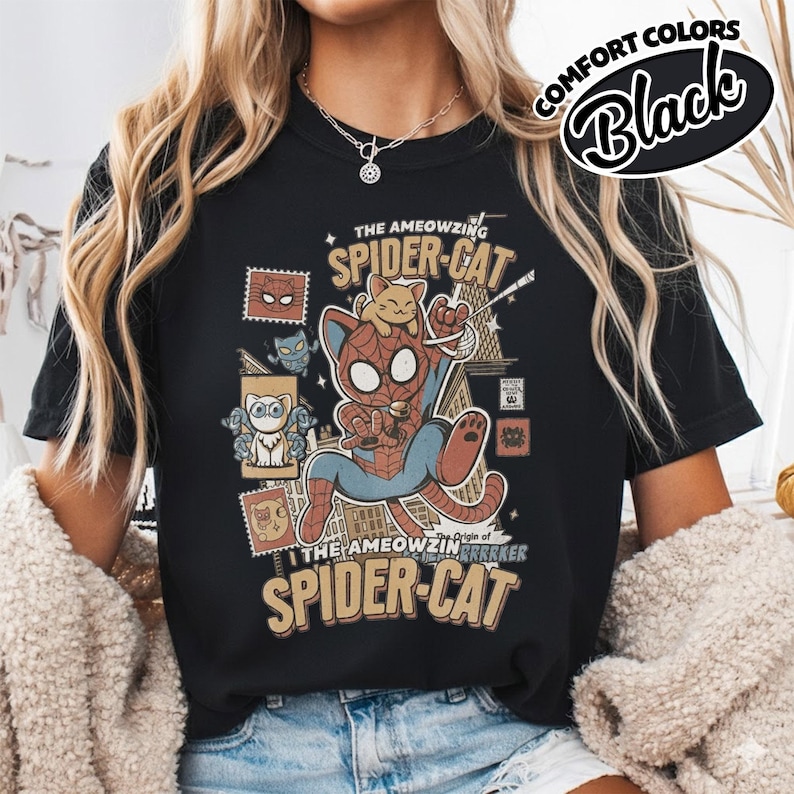 May include: Black t-shirt featuring a cartoon illustration of a cat dressed as Spider-Man, with the text "THE AMEOWZING SPIDER-CAT." The design includes a smaller cat character and various stamp-like graphics. The words "COMFORT COLORS" and "Black" are also visible.