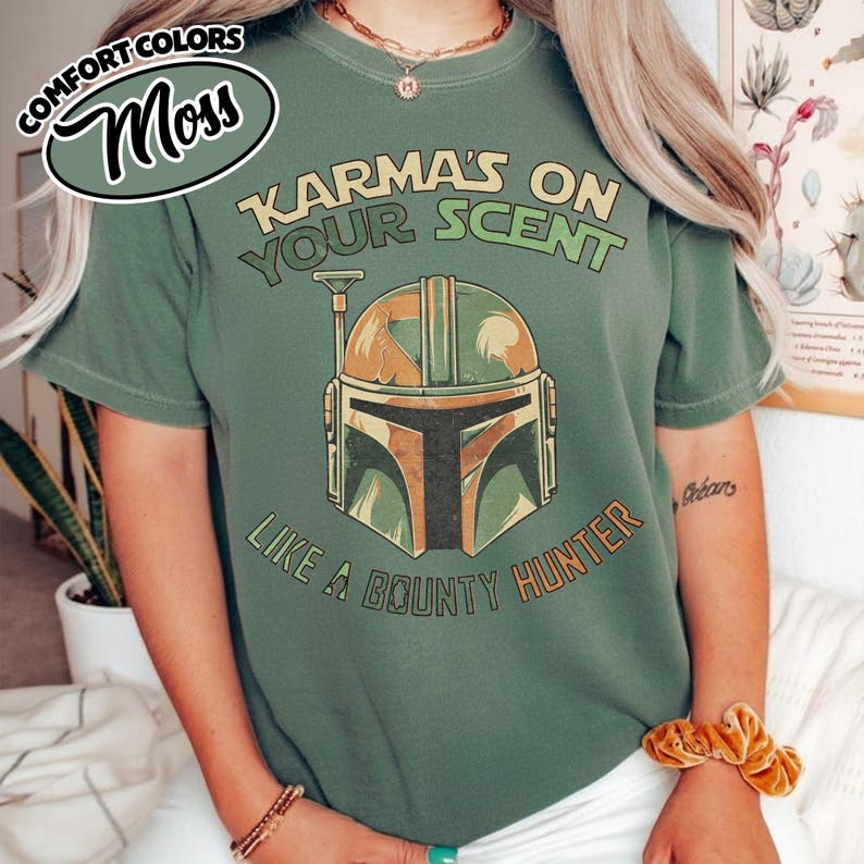 May include: Moss green t-shirt with the text "Karma's on Your Scent Like a Bounty Hunter" and a graphic of a Mandalorian helmet in shades of green, brown, and orange. The shirt has short sleeves.