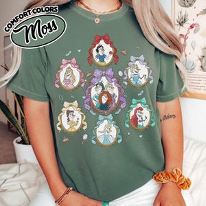 May include: Moss green t-shirt featuring framed illustrations of animated characters. The illustrations are surrounded by decorative bows. The top left corner of the shirt has the text "COMFORT COLORS" and "Moss" in a circular design.