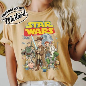 May include: Mustard-colored t-shirt featuring a Star Wars graphic with characters from Toy Story. The design includes Woody as a Jedi, Buzz Lightyear, and other characters. The words "COMFORT COLORS" and "Mustard" are printed on the top left.