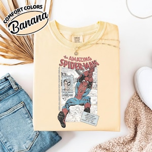 K&ouml;nnte beinhalten: Hellgelbes T-Shirt mit einer Grafik von Spider-Man in Aktion, mit dem Text "The Amazing Spider-Man". Das Shirt tr&auml;gt auch die Aufschrift "Comfort Colors" und "Banana".