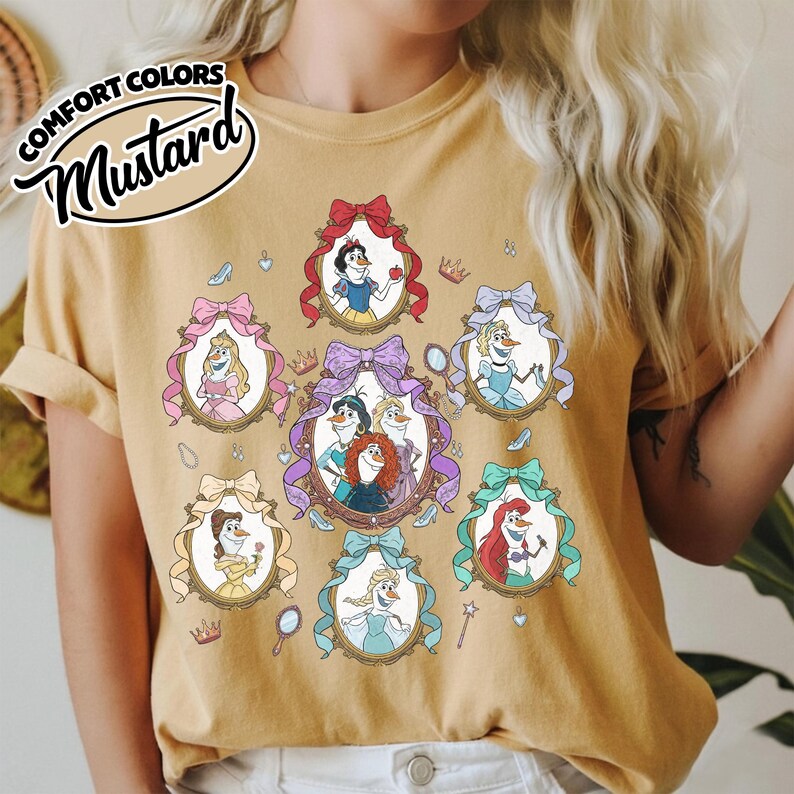 May include: Mustard-colored t-shirt featuring framed illustrations of animated princesses. Each frame is adorned with a bow. The shirt includes the text "COMFORT COLORS" and "Mustard" in a stylized font.
