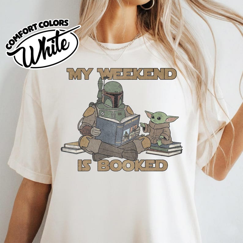 Może przedstawiać: Biała koszulka Comfort Colors z grafiką Boba Fetta czytającego książkę Grogu, z napisem "MY WEEKEND IS BOOKED". Projekt wykorzystuje kolory brązowy, zielony i beżowy. Koszulka ma okrągły dekolt i kr&oacute;tkie rękawy.