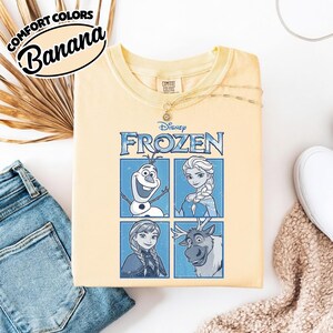 May include: A light yellow sweatshirt featuring the word "Frozen" and images of characters Olaf, Elsa, Anna, and Sven. The sweatshirt has the words "Comfort Colors" and "Banana" in a black oval on the top left.
