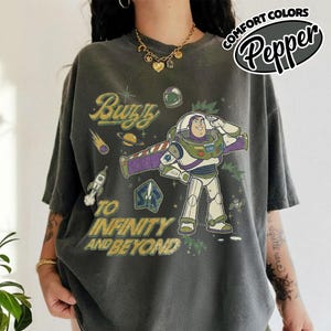 May include: A charcoal gray short-sleeve t-shirt featuring a graphic of Buzz Lightyear saluting. The design includes the text Buzz and To Infinity and Beyond, along with illustrations of a rocket, a planet, and stars in yellow and green tones.