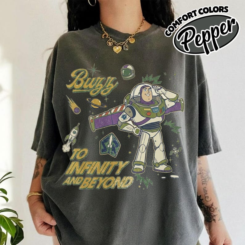 May include: A charcoal gray short-sleeve t-shirt featuring a graphic of Buzz Lightyear saluting. The design includes the text Buzz and To Infinity and Beyond, along with illustrations of a rocket, a planet, and stars in yellow and green tones.