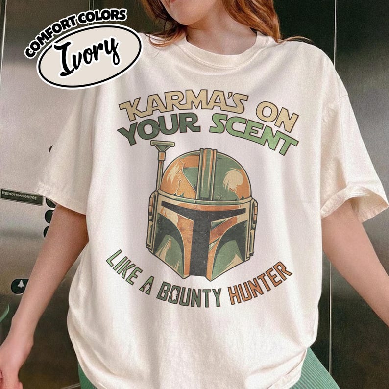 May include: Ivory-colored t-shirt with the text "Karma's on Your Scent" and "Like a Bounty Hunter." The graphic features a Mandalorian helmet in shades of green, orange, and brown. The shirt is a Comfort Colors brand.