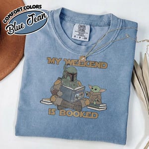 Może przedstawiać: Niebieska koszulka Comfort Colors z napisem "MY WEEKEND IS BOOKED" i grafiką Boba Fetta czytającego Grogu. Koszulka ma okrągły dekolt i kr&oacute;tkie rękawy. Złoty naszyjnik jest zarzucony na koszulkę.